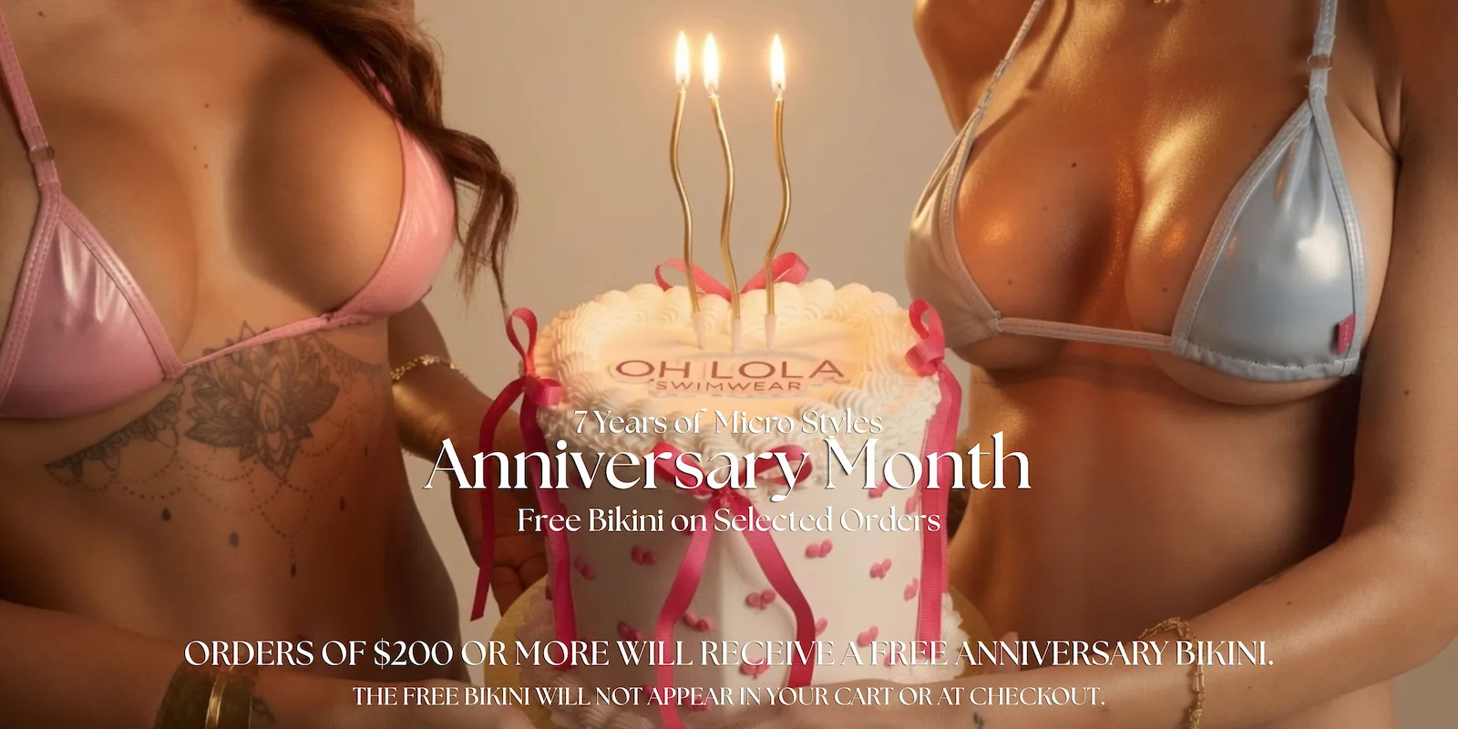 Two models wearing Oh Lola Swimwear pink and light blue micro bikinis holding a white birthday cake decorated with pink hearts, ribbons, and the Oh Lola Swimwear logo, topped with three lit gold candles in a warm golden studio editorial photoshoot.